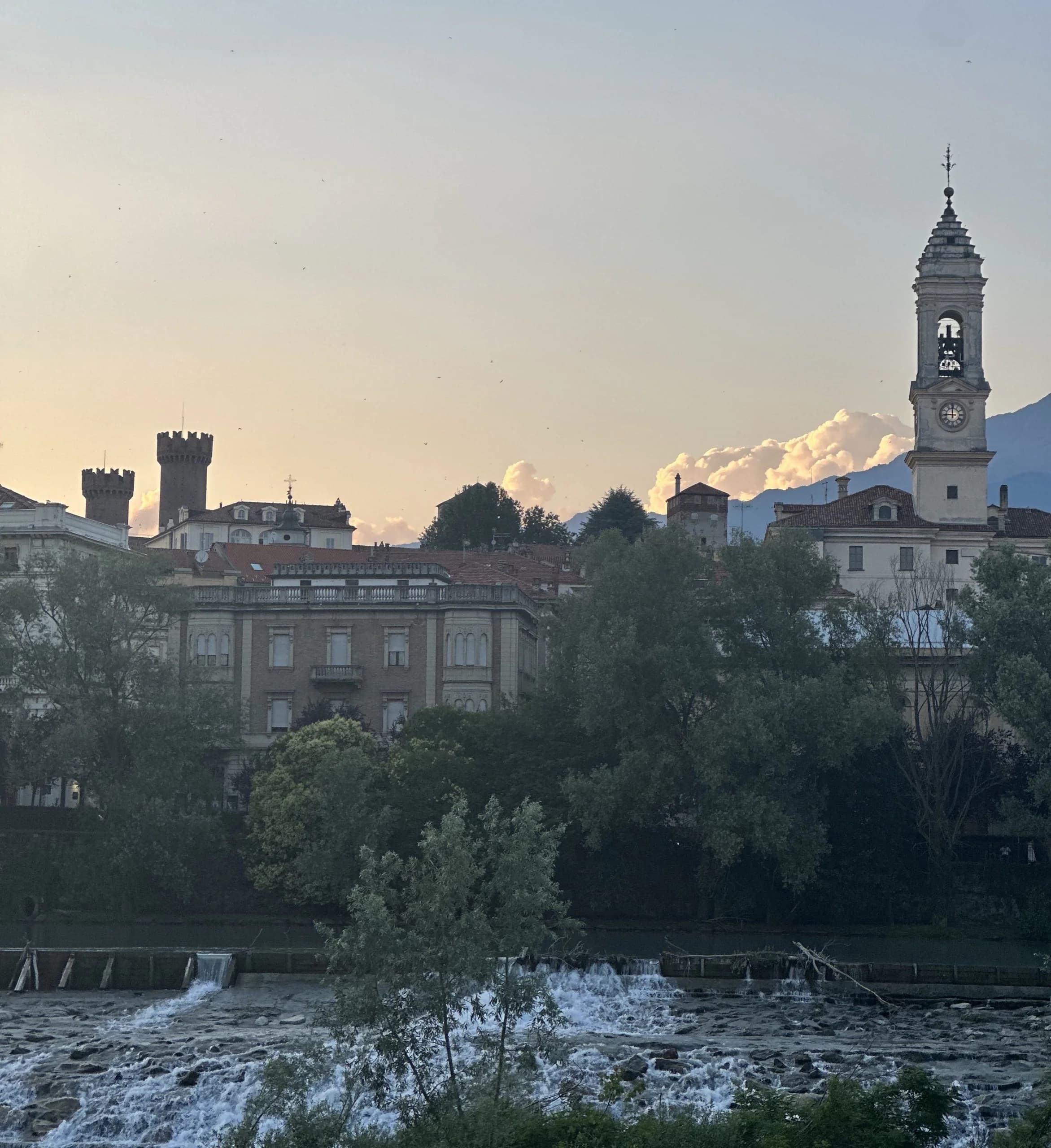 Vista do rio ao entardecer: a torre de um castelo e o campanário de uma igreja acima do casario, as árvores da margem e a corredeira sobre as pedras em primeiro plano